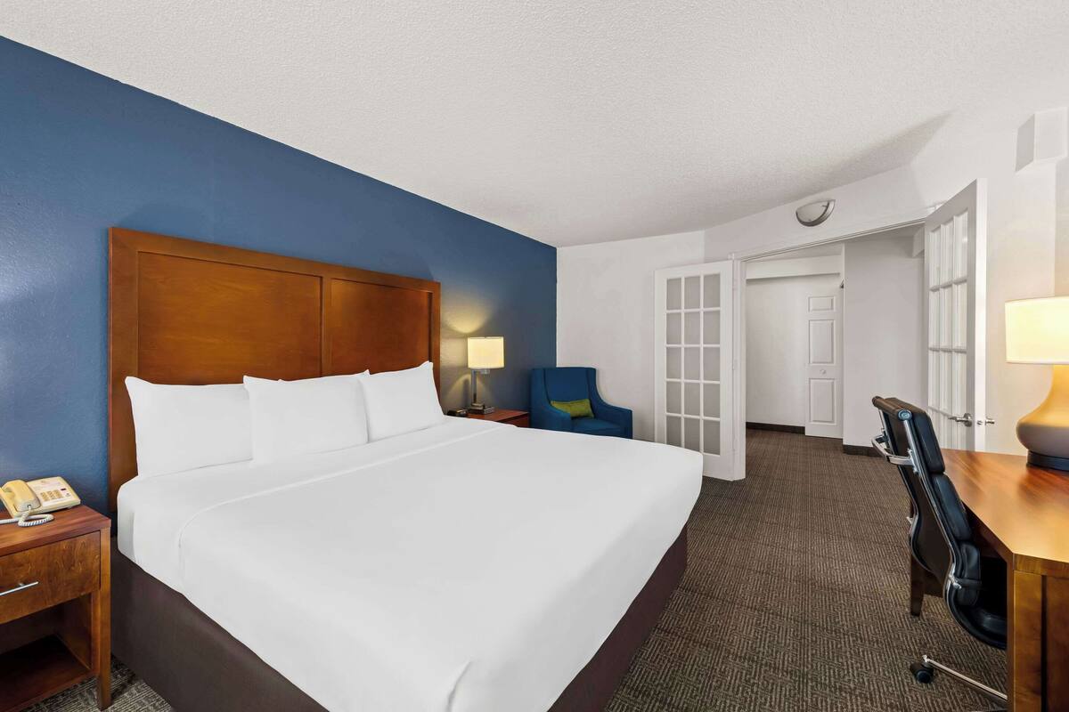 A hotel room with a large bed, a wooden headboard, a desk with a lamp, a blue chair, and a phone on the nightstand.
