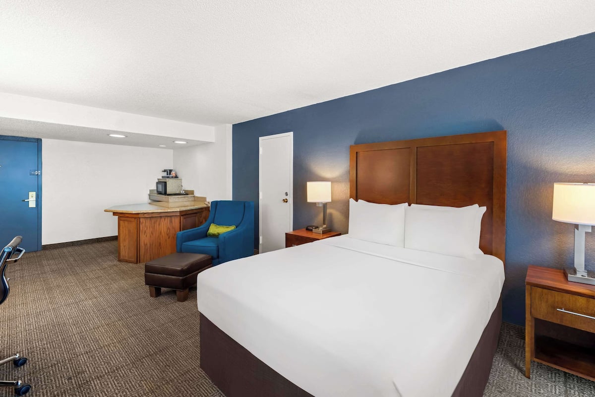 A hotel room with a large bed, a desk with a chair, a lamp, and a small table.