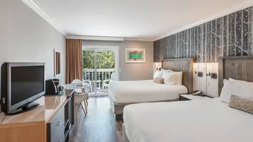 Habitación de hotel con dos camas, televisión, escritorio y balcón con vista a árboles.