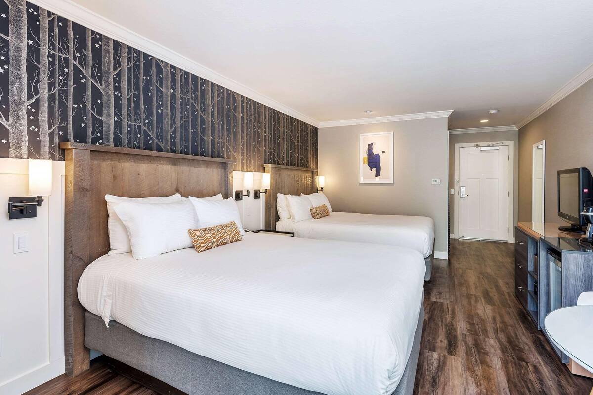 A hotel room with two beds, a wooden headboard, a wall-mounted TV, and a small desk area.