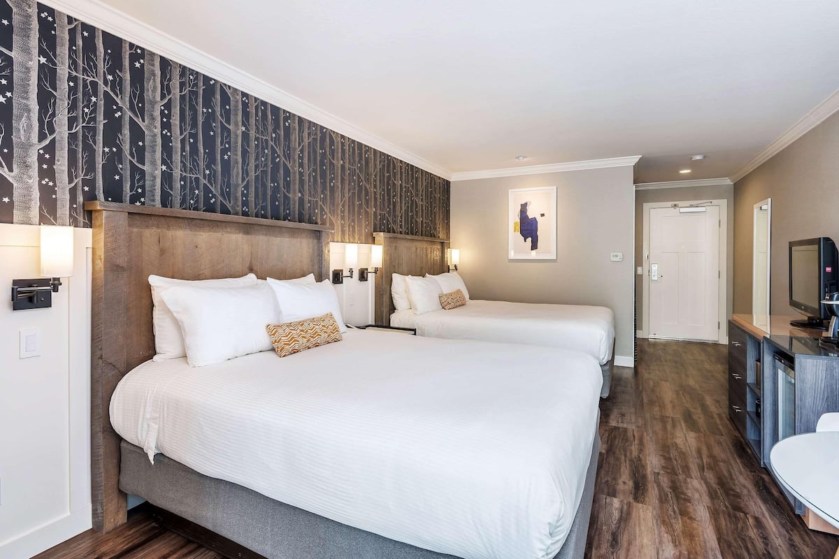 A hotel room with two beds, a wooden headboard, a wall-mounted TV, and a small desk area.