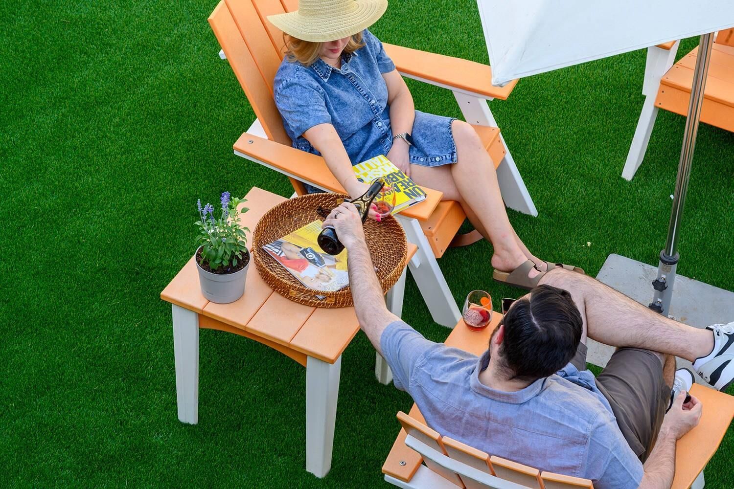 Dos personas sentadas en sillas al aire libre, con una mesa entre ellas, una planta en maceta y una revista.