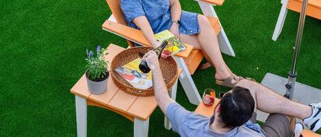 Dos personas sentadas en sillas al aire libre, con una mesa entre ellas, una planta en maceta y una revista.