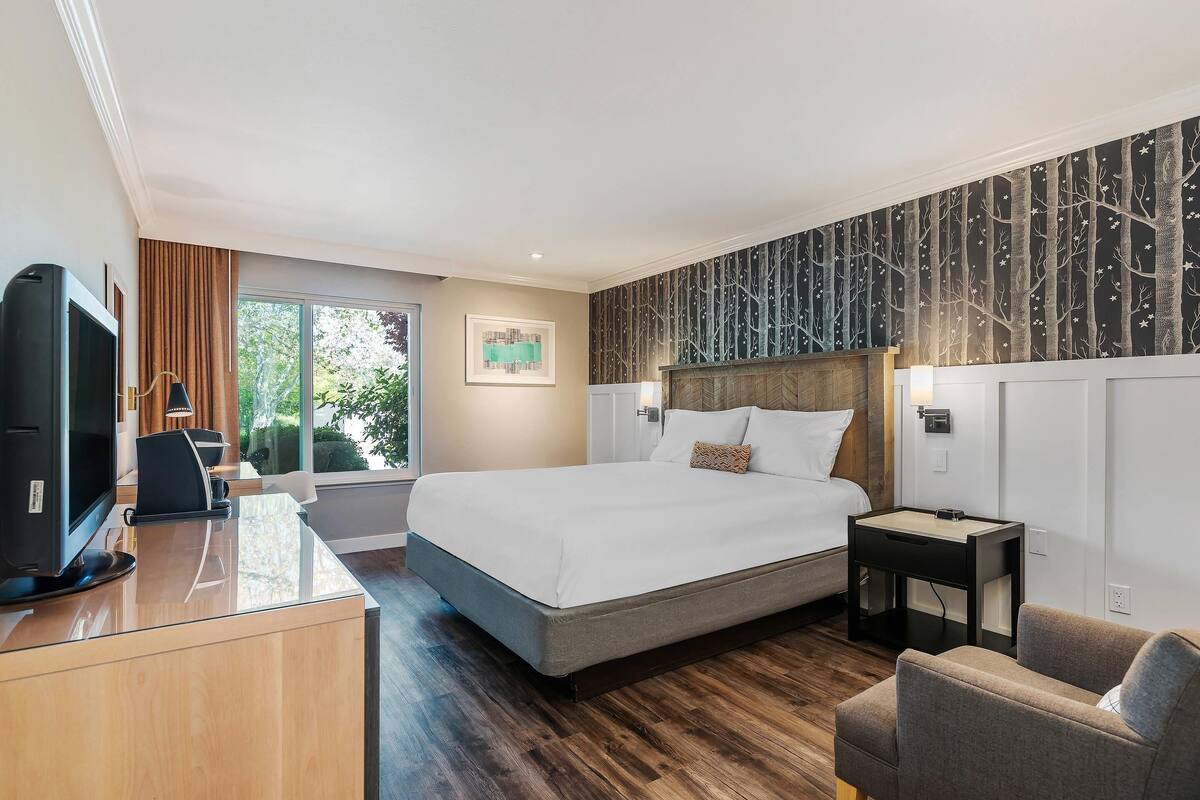 A modern hotel room with a large bed, a desk with a computer, a chair, and a small table.