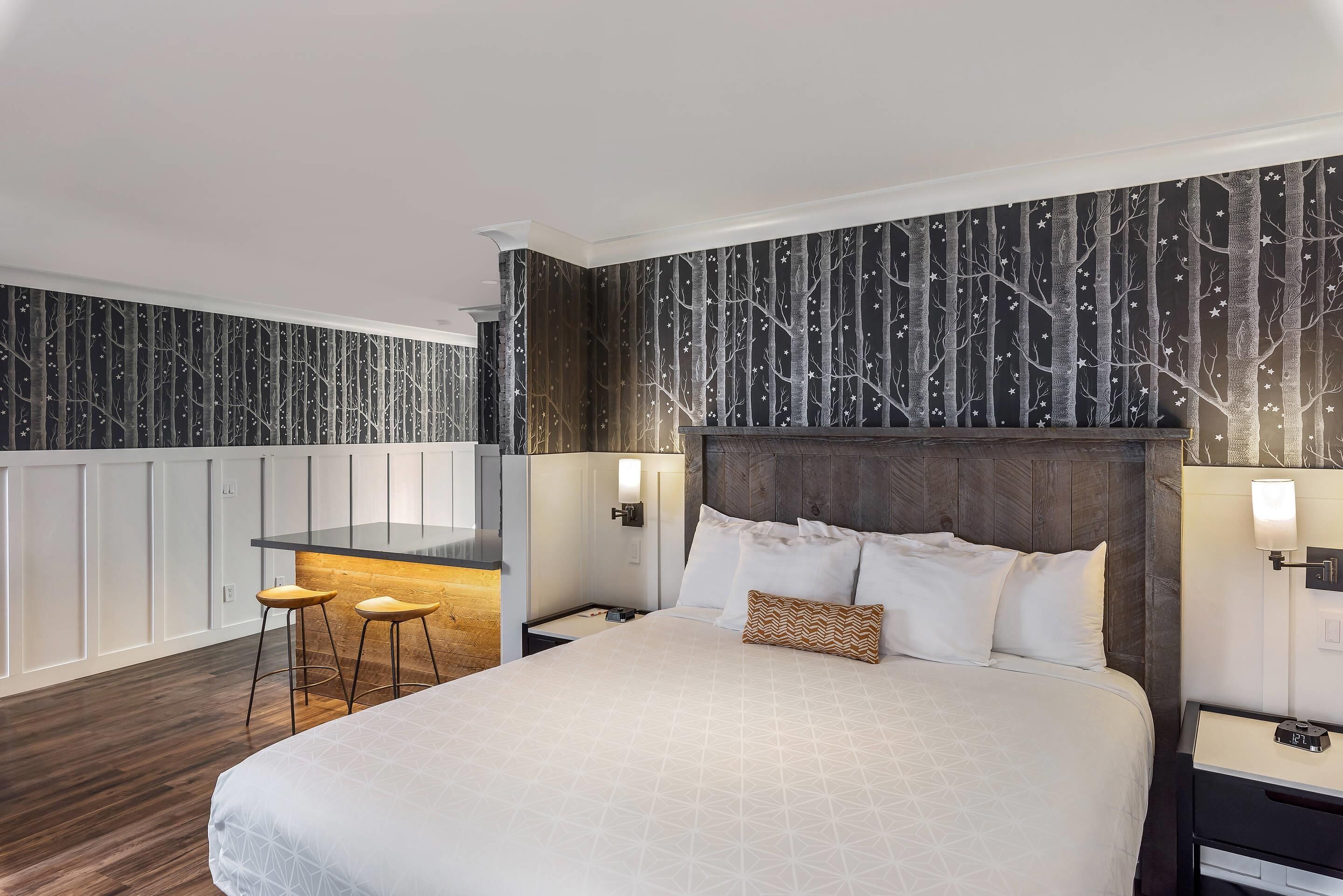 A modern hotel room with a large bed, a small kitchen area, and a seating space with stools.