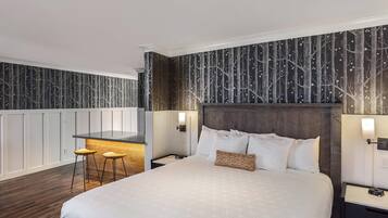 A modern hotel room with a large bed, a small kitchen area, and a seating space with stools.