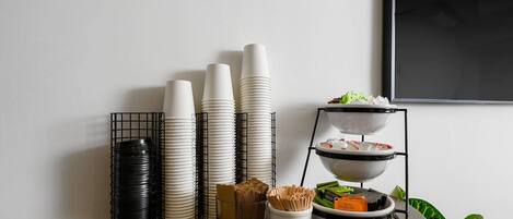 A buffet setup with stacked plates and bowls on a table, a tray with food items, and a plant in the corner.