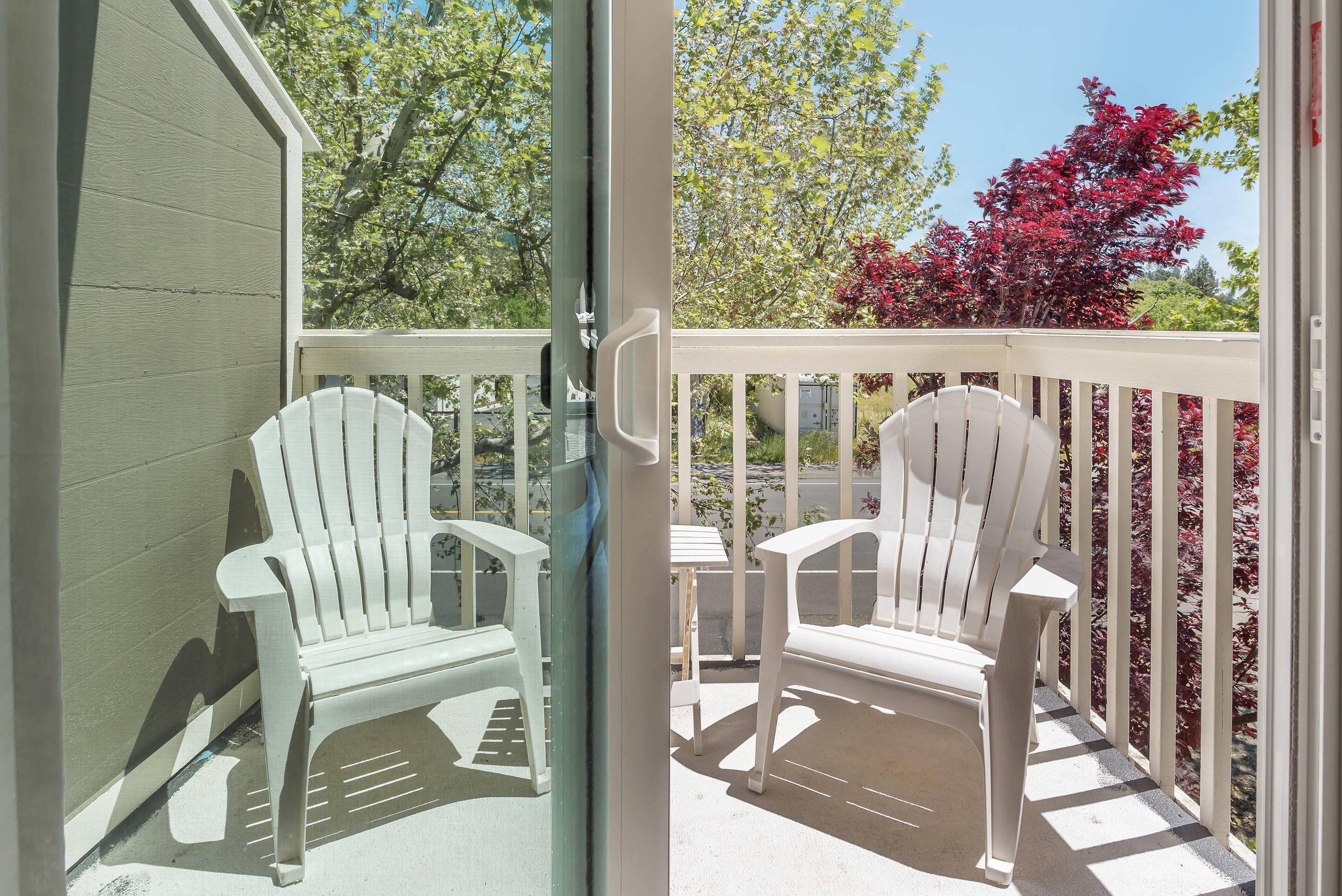 A balcony with two white Adirondack chairs, a small table, and a view of trees and foliage.