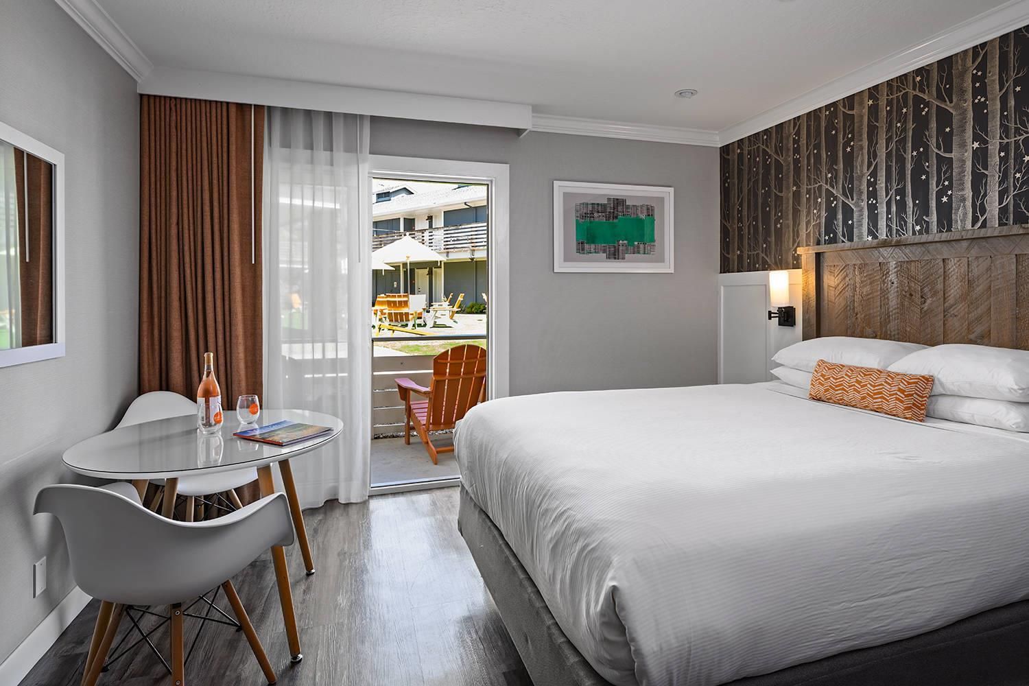 A hotel room with a bed, a table with chairs, a balcony with a view of buildings, and a wall with a cityscape painting.