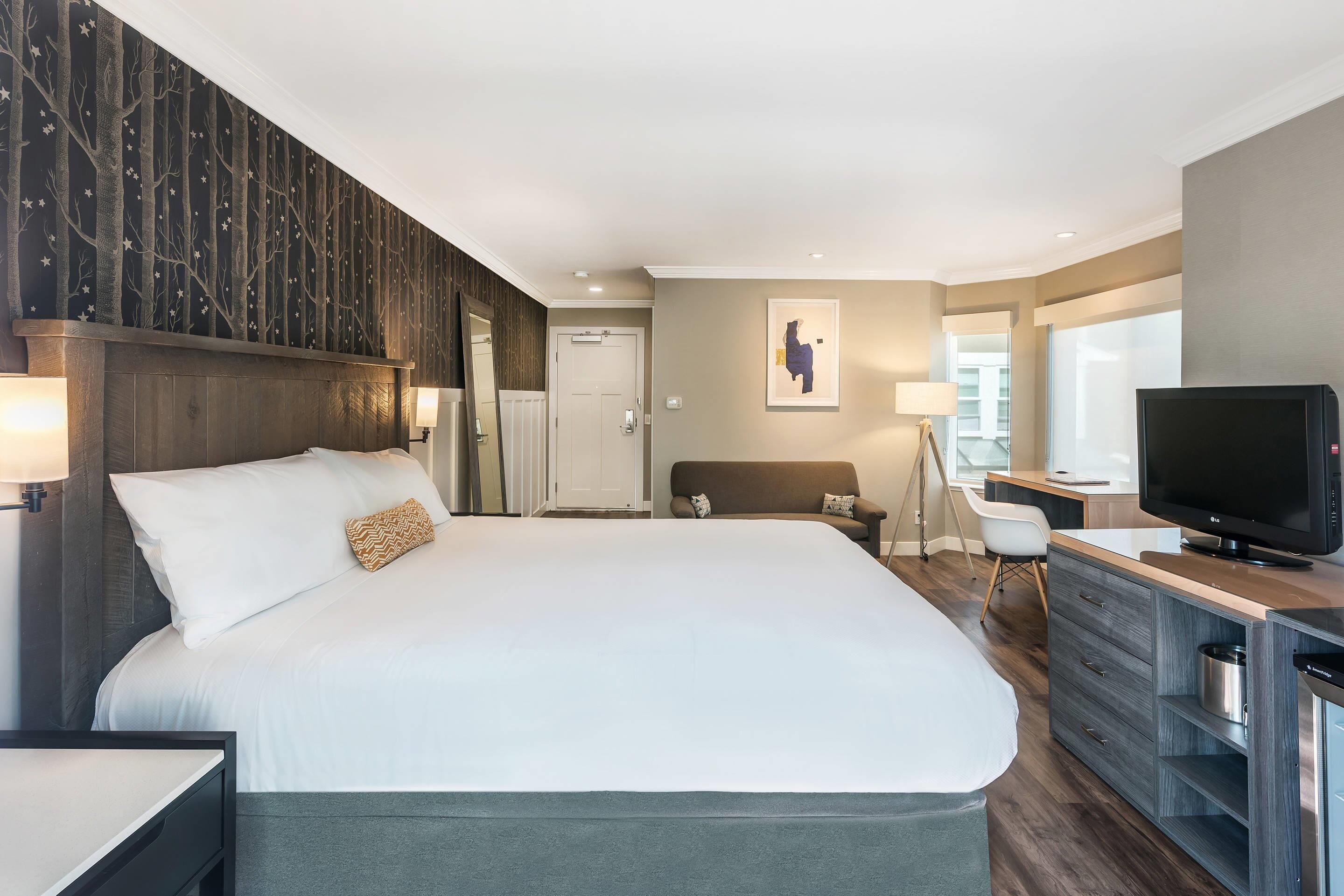 A modern hotel room with a large bed, a desk with a TV, and a view of the city.