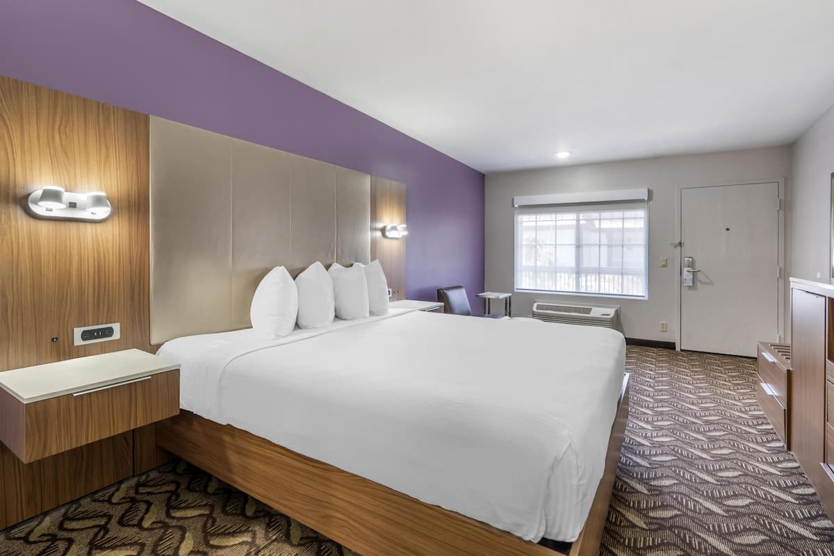 Ein Hotelzimmer mit einem großen Bett, Nachttischen, einem Schreibtisch, einem Fenster mit Jalousien und einer Tür.