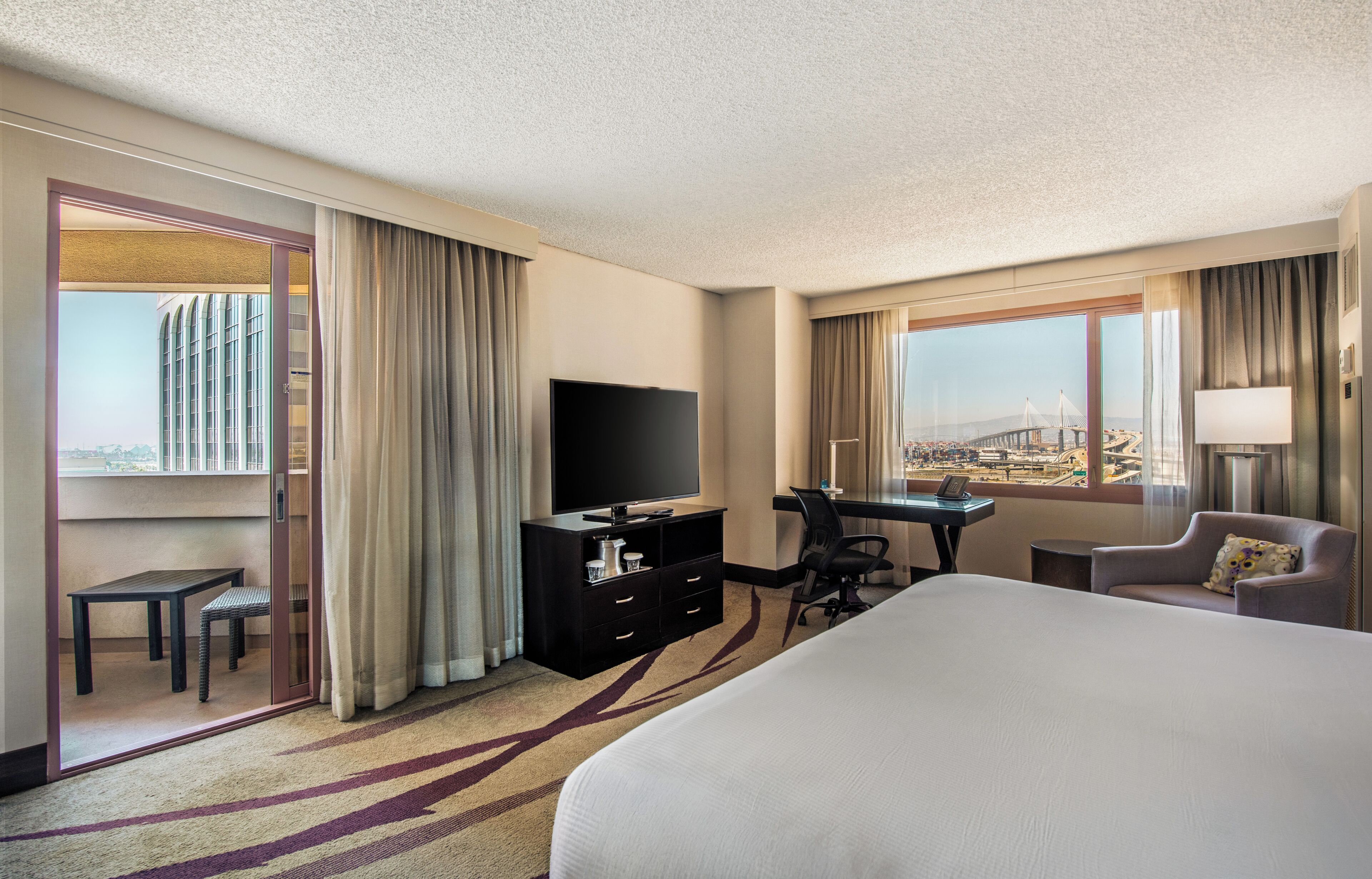 A hotel room with a bed, a desk, a television, a chair, and a view of a bridge.