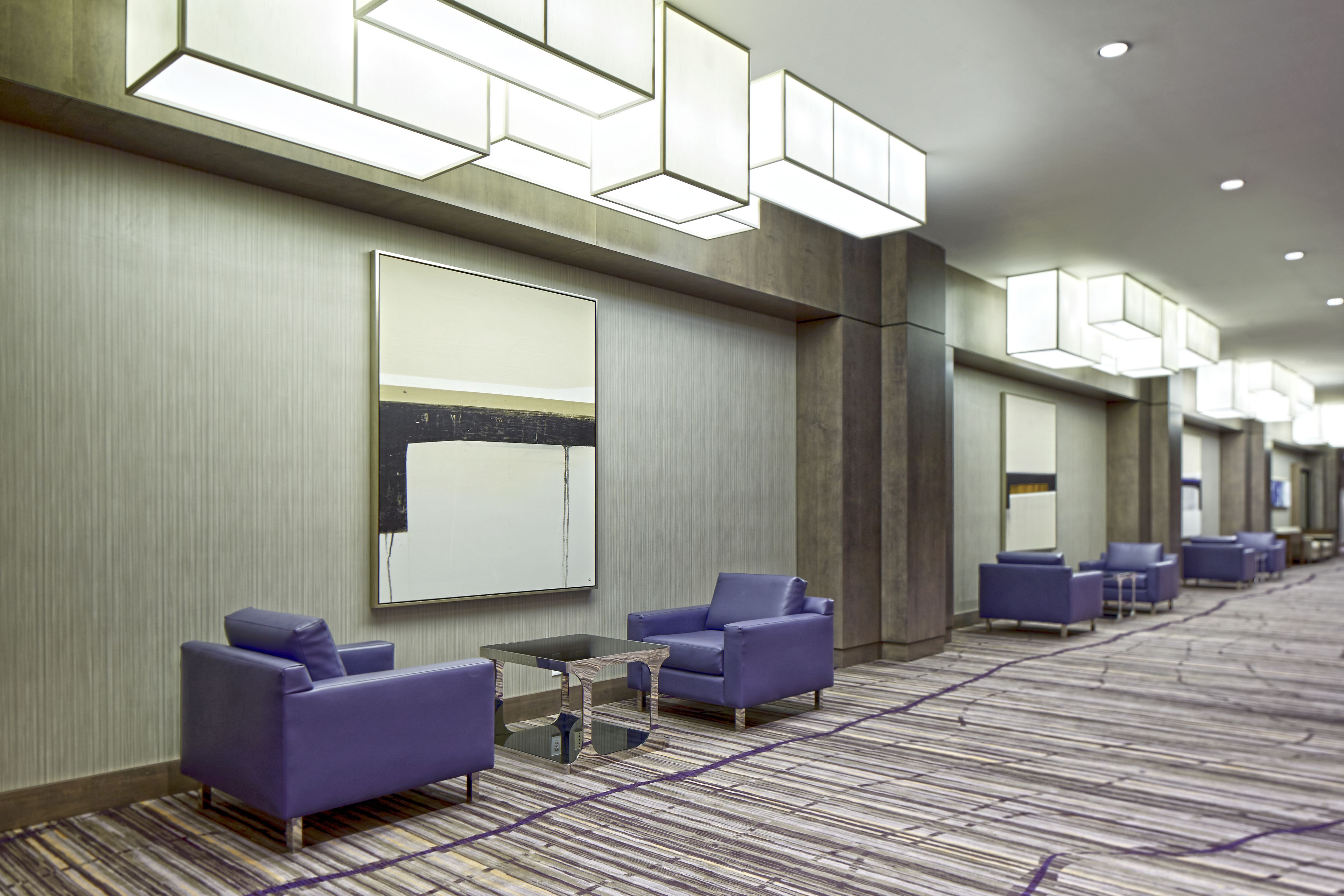 Eine Hotte lobby mit einer langen Korridor, violette Sitzgelegenheiten, einem großen Wandbildschirm und geometrischen Deckenleuchten.