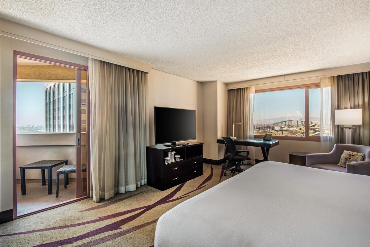 Habitación de hotel con una cama grande, un escritorio con una silla, televisión y vista a un puente.