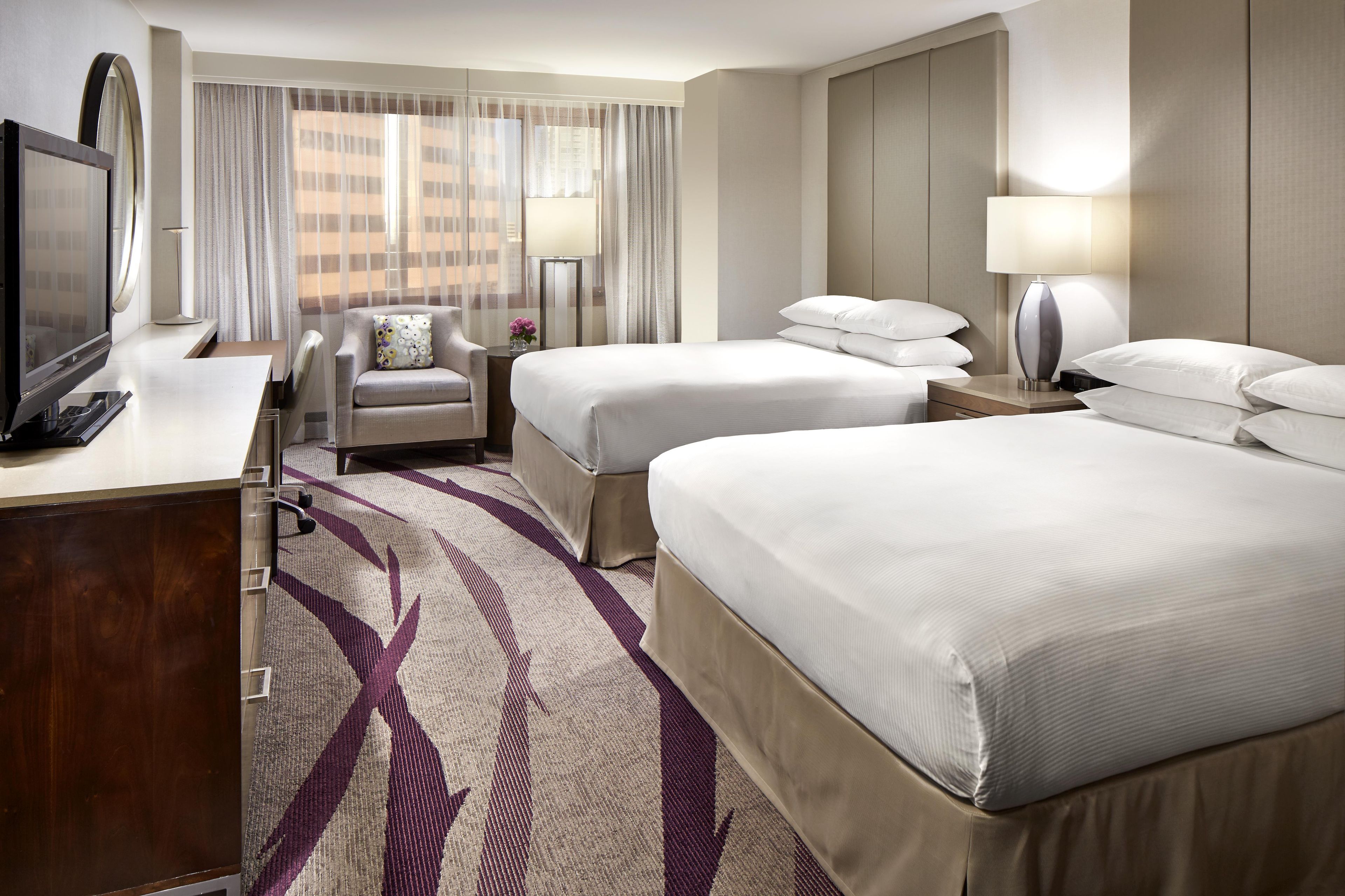 A hotel room with two beds, a desk with a TV, a chair, a lamp, and a view of buildings through sheer curtains.
