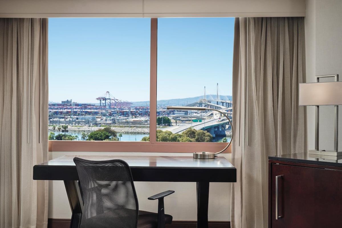 A hotel room with a desk, chair, and a view of a cityscape with a bridge and port.