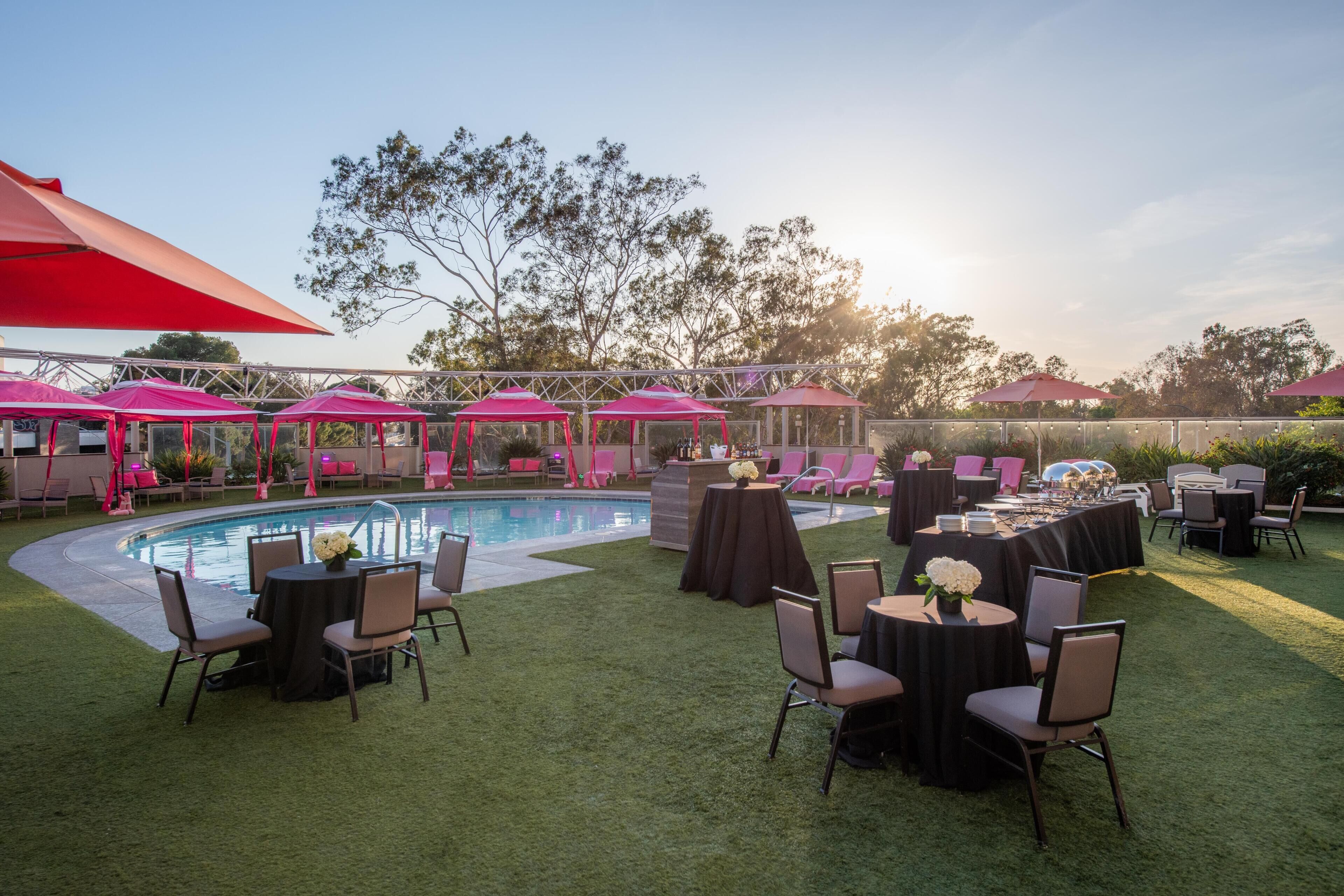 An outdoor pool area with multiple tables and chairs, surrounded by pink umbrellas and a clear sky.