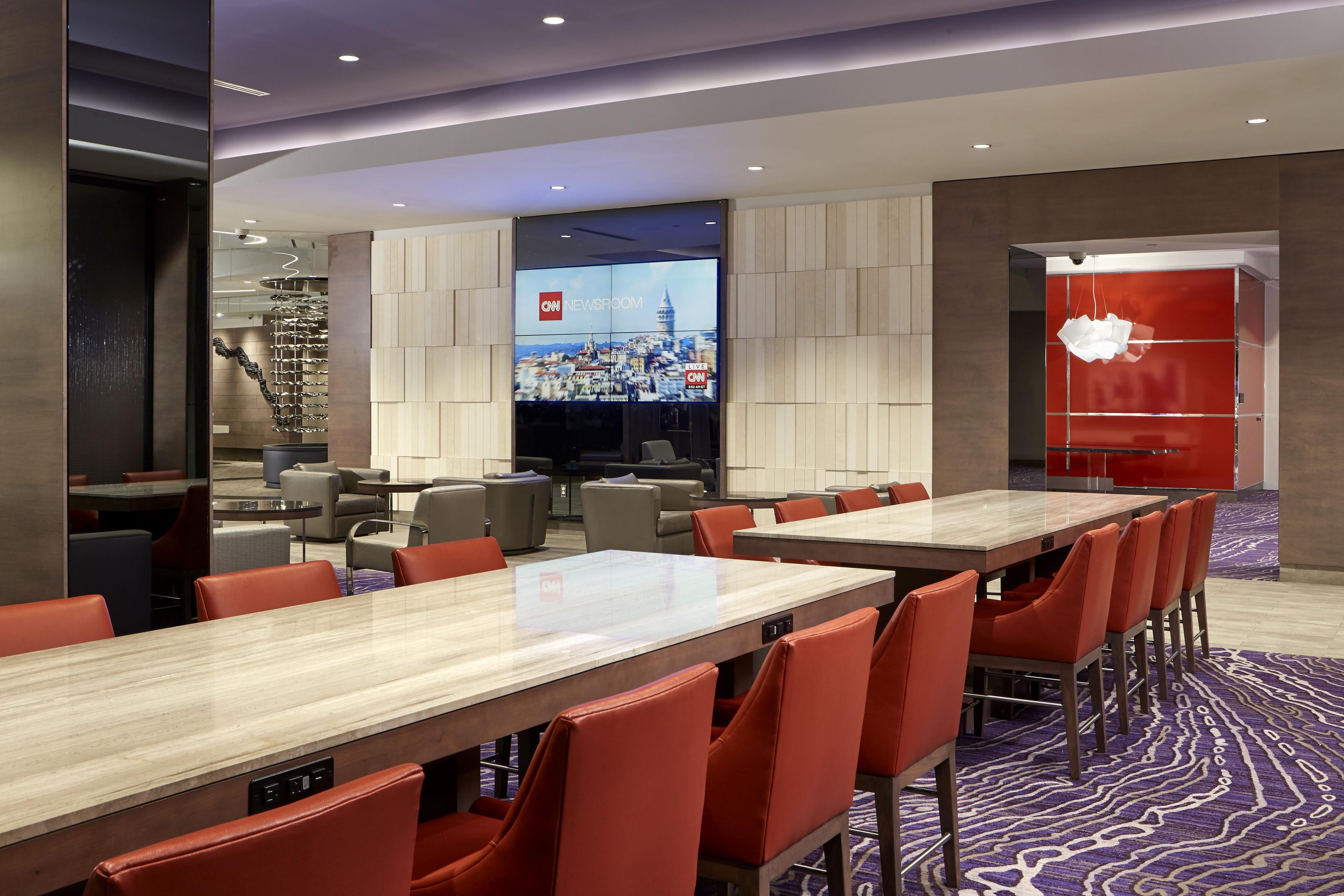 A modern hotel lobby with a CNN Newsroom display, seating areas, and a staircase.