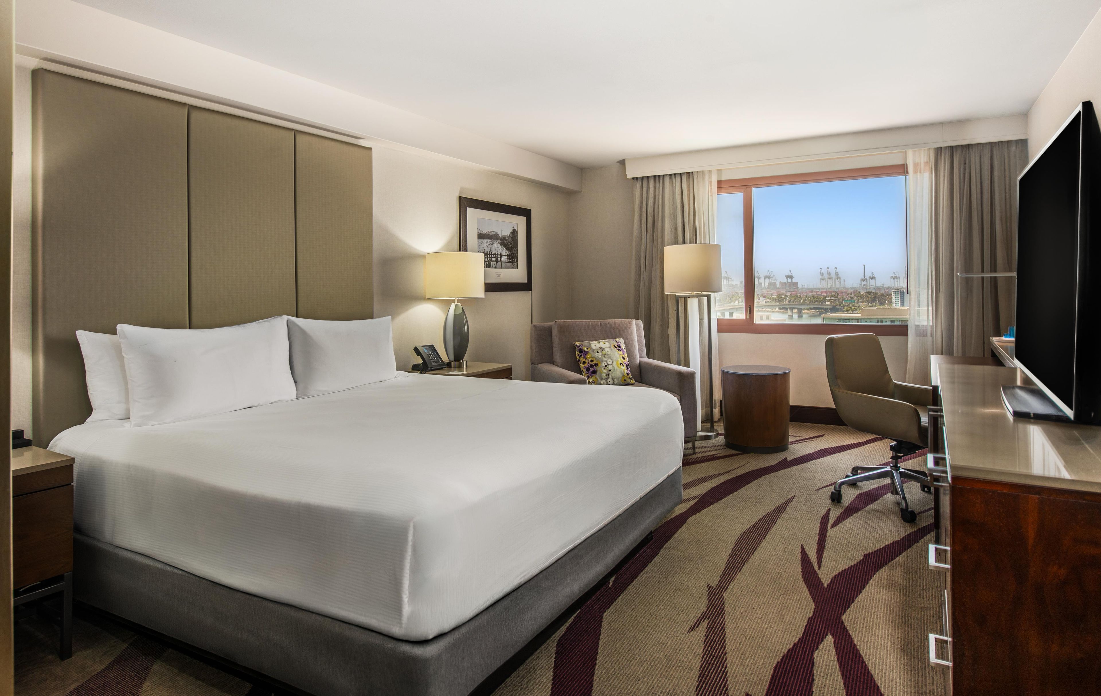Habitación de hotel con una cama grande, un escritorio, una silla, un televisor y vistas a la ciudad.