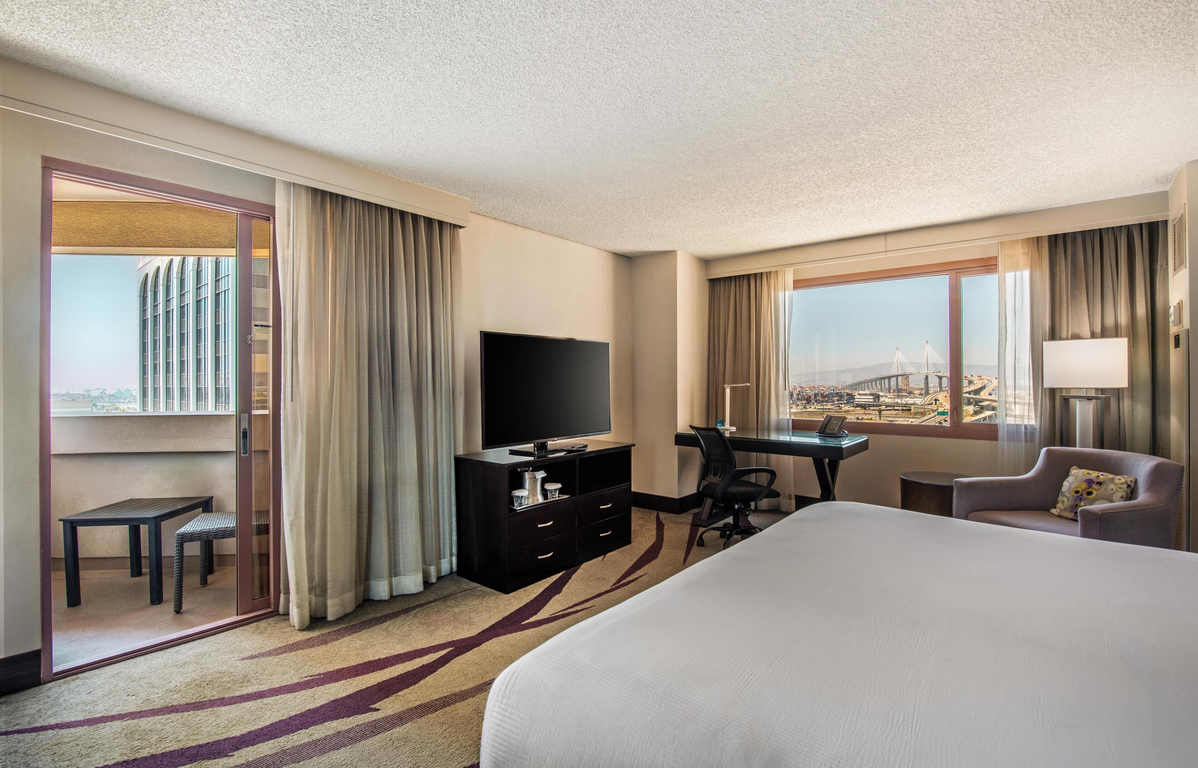 A hotel room with a large bed, a desk with a chair, a television, and a view of a bridge.