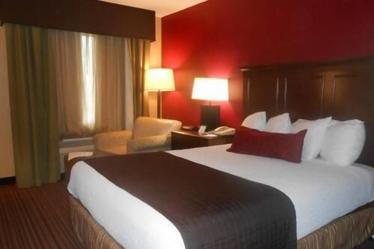 Quarto de hotel com uma cama grande, uma cadeira, mesa de cabeceira com abajur e janela com cortinas.