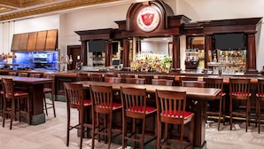 A bar area with multiple wooden tables and chairs, a bar counter with a variety of bottles, and a logo above the counter.