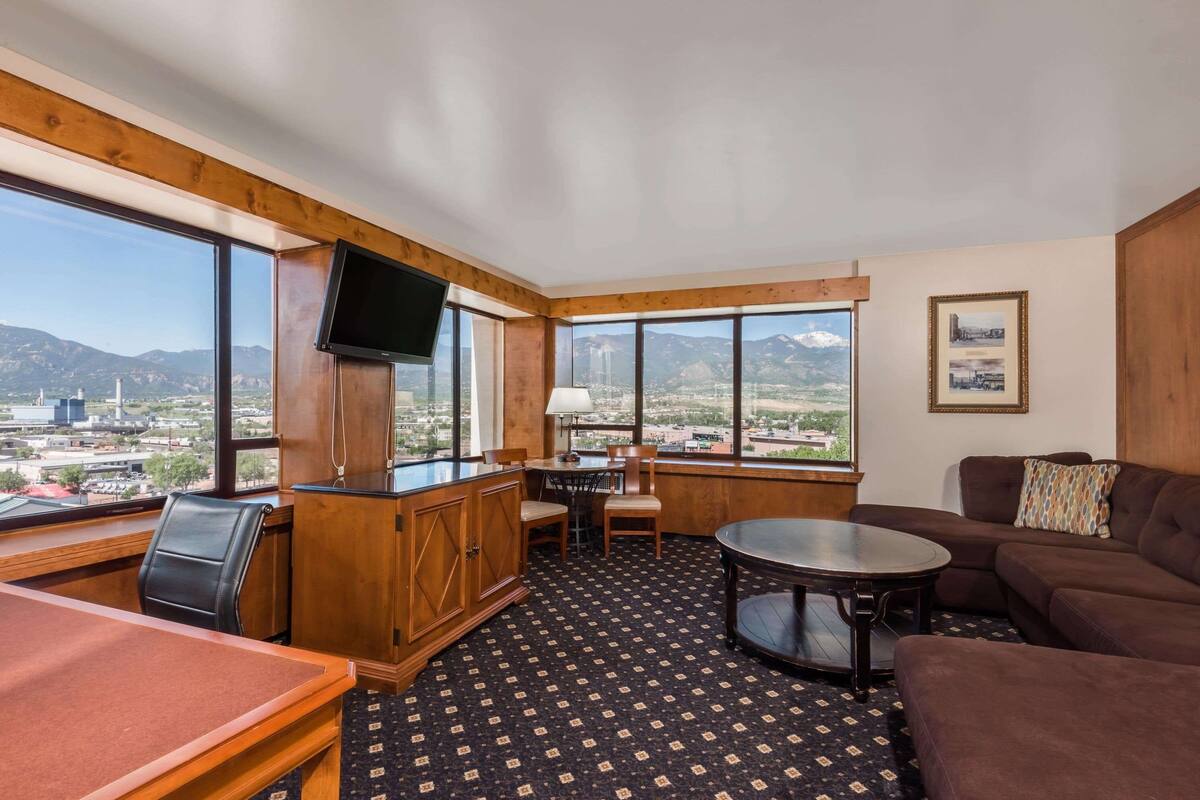 A hotel room with a large window offering a mountain view, a wooden desk, a sofa, and a television mounted on the wall.