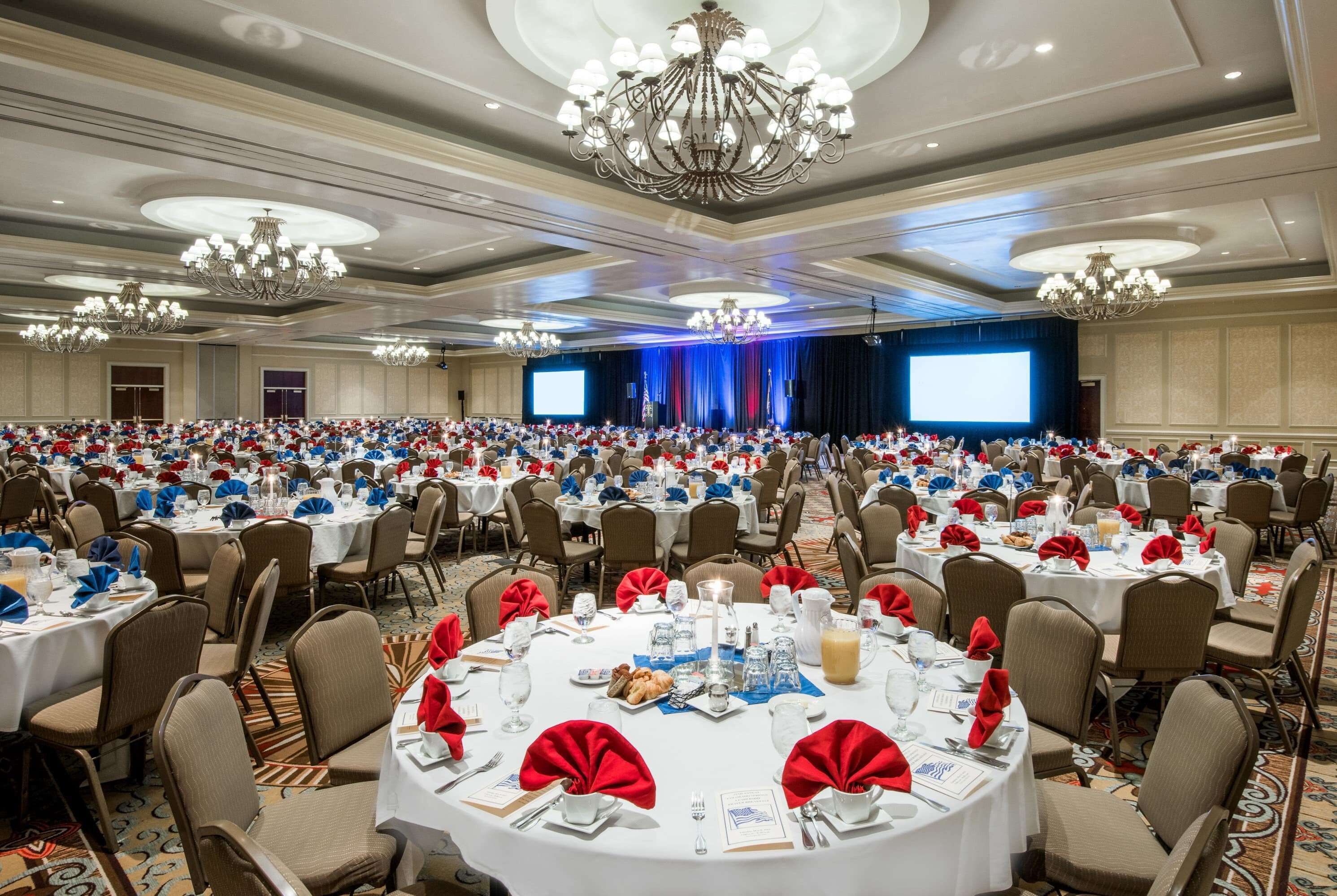 A large banquet hall with round tables set for a formal event, featuring chandeliers and a stage with a blue backdrop.