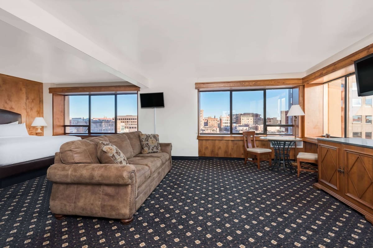 A hotel room with a large bed, a sofa, a dining area with chairs, a TV, and large windows offering a city view.