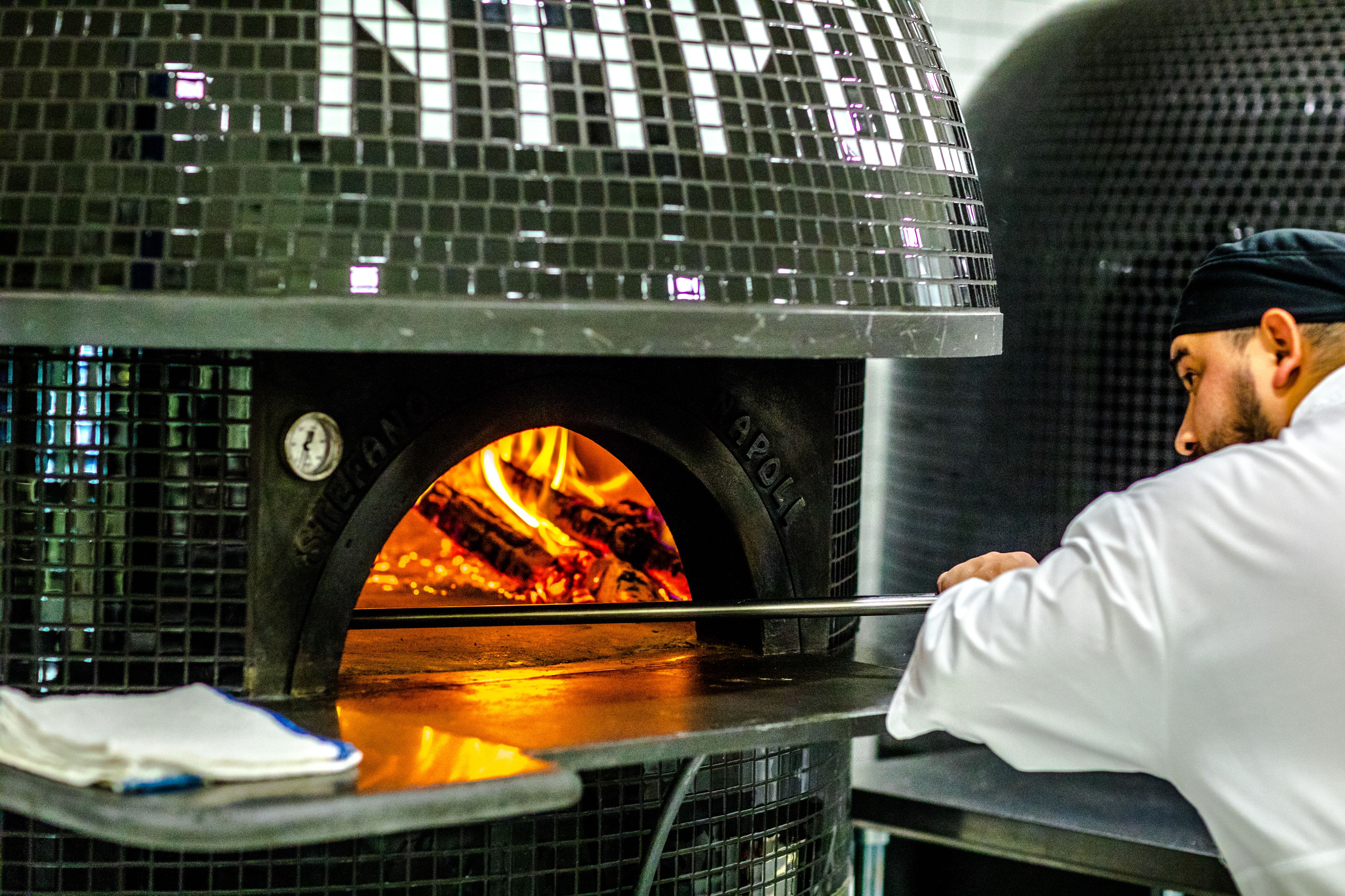 Ein Pizzaofen mit Feuer darin, eine Person in einem weißen Kittel und eine Kachelwand.