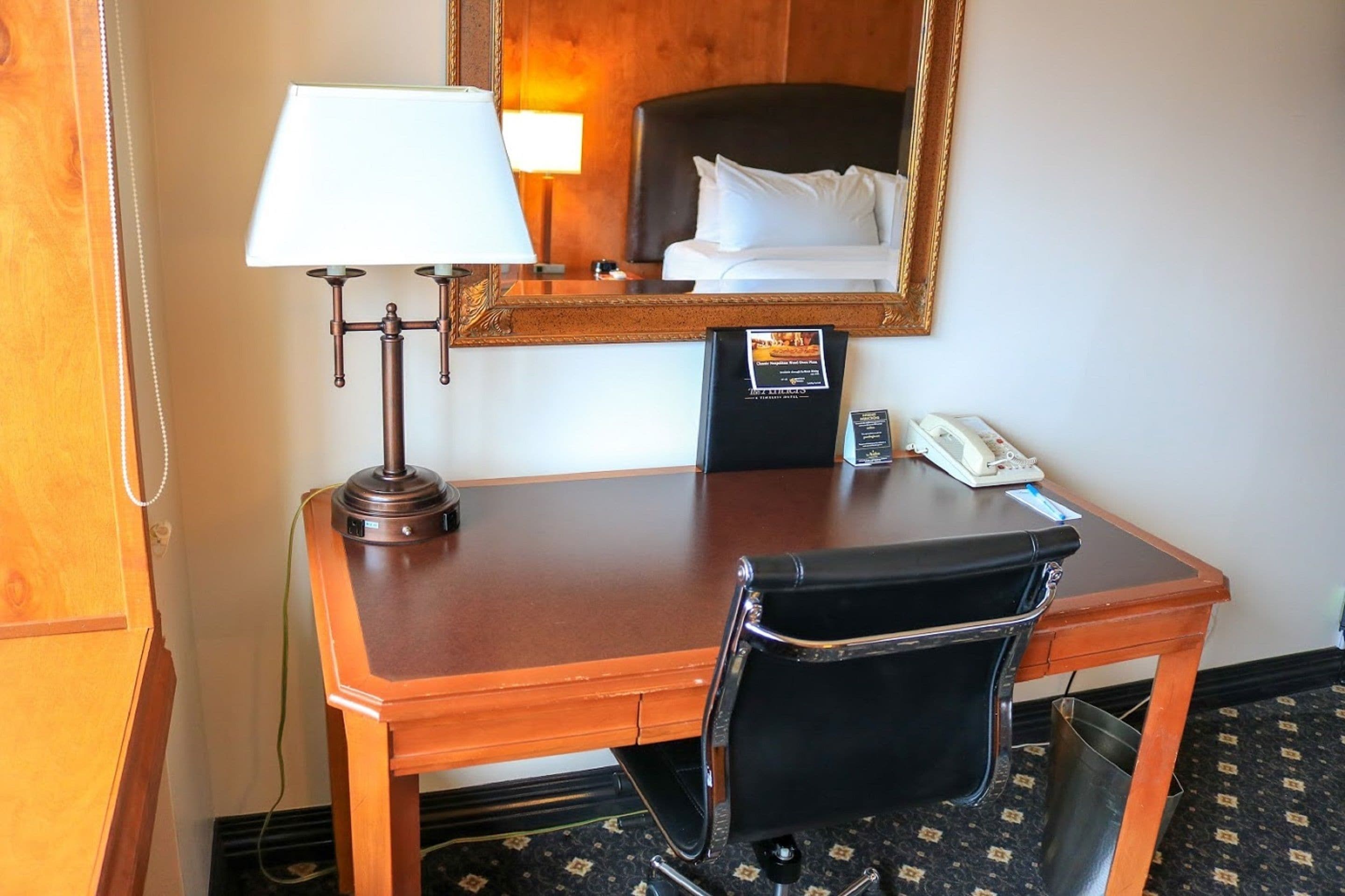 Habitación de hotel con un escritorio de madera, una silla de cuero negro, una lámpara, una cama con sábanas blancas, una mesita de noche con un teléfono y un espejo en la pared.