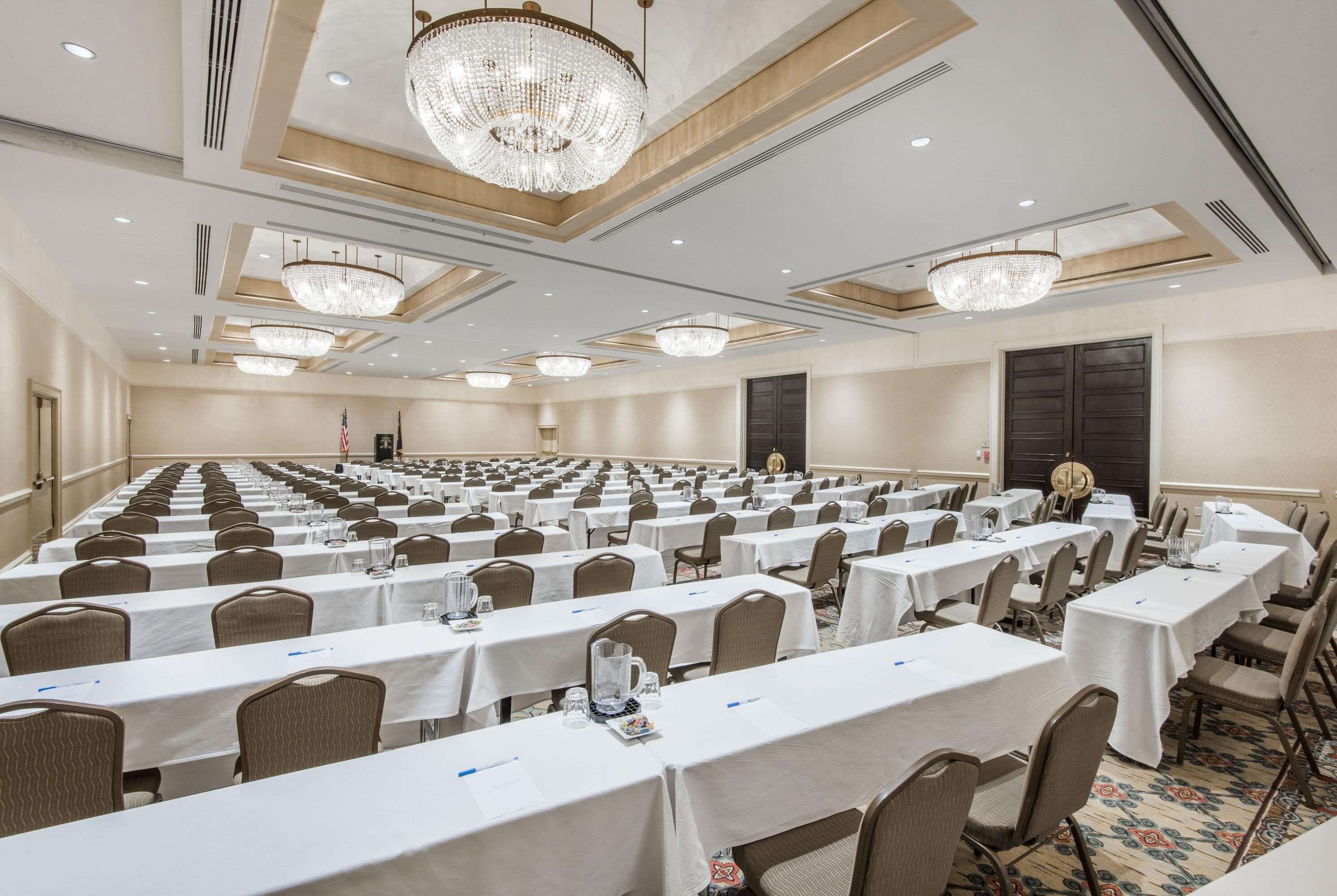 A large conference room with rows of tables and chairs, chandeliers, and a flag.