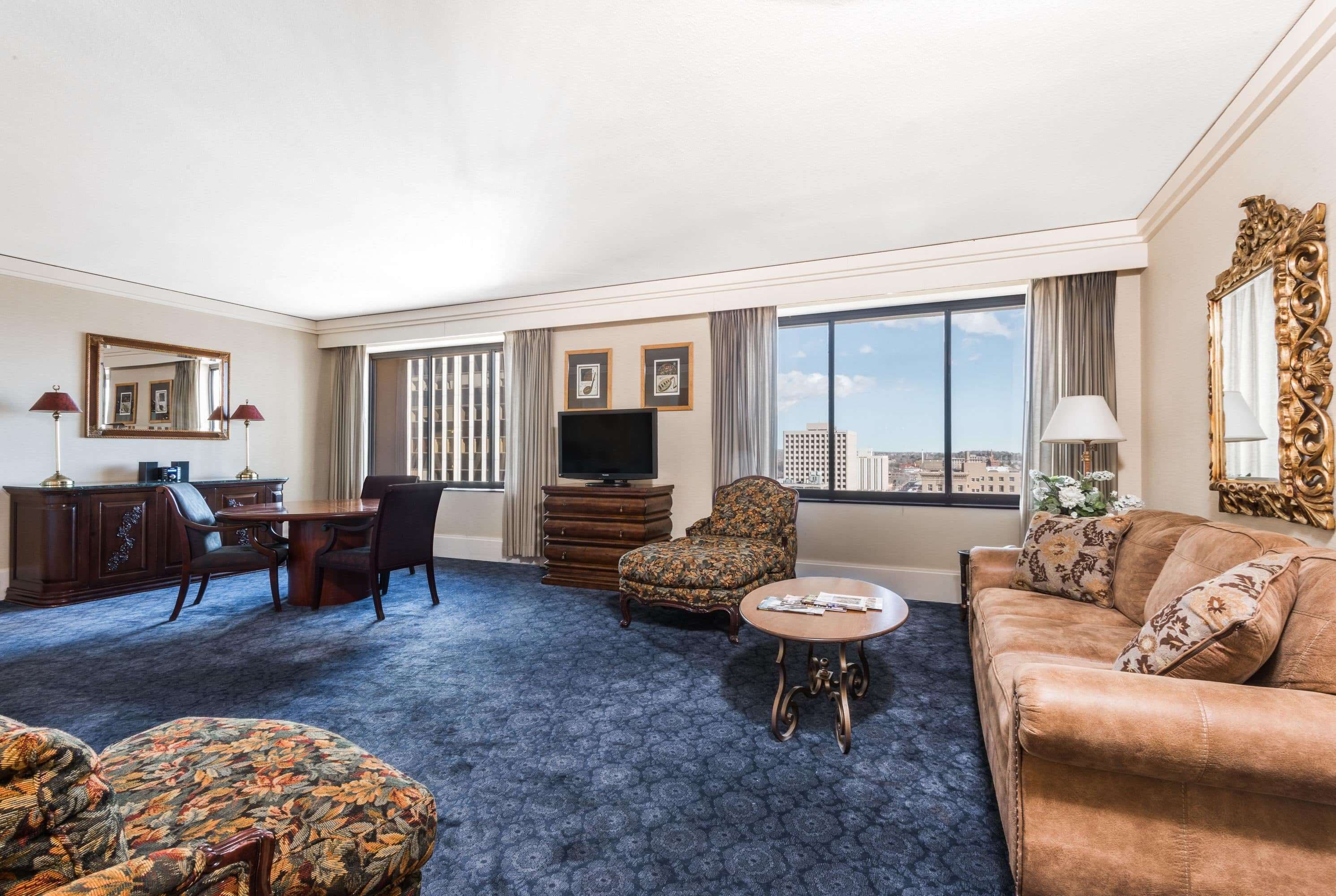 A hotel room with a large window, a flat-screen TV, a dining area with a table and chairs, a sofa, and a patterned armchair.