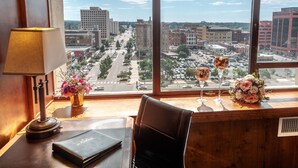 A hotel room with a large window offering a city view, a desk with a lamp, a vase with flowers, and a glass of wine.