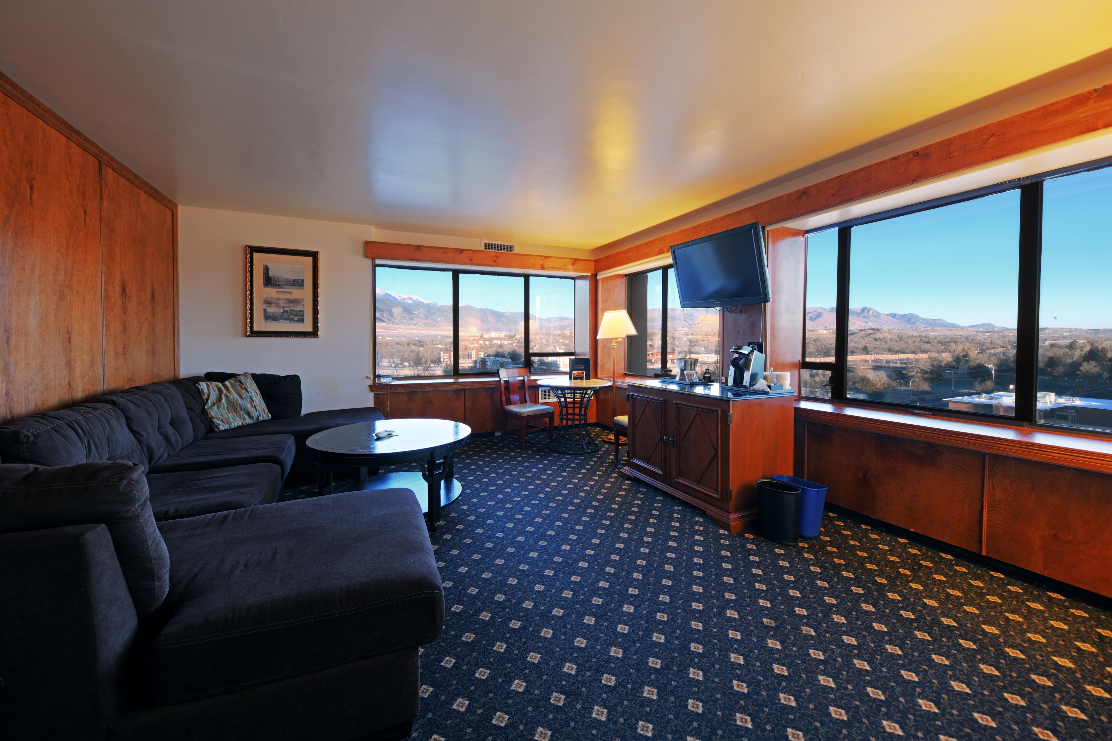 A hotel room with a dark-colored sofa, a small round table, a TV, and a view of mountains through large windows.