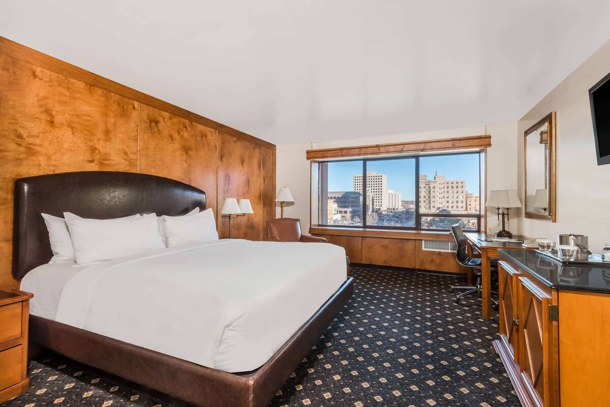A hotel room with a large bed, a desk, a chair, a window with a city view, and a wooden headboard.