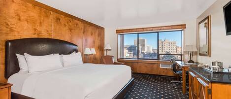 A hotel room with a large bed, a desk, a chair, a window with a city view, and a wooden headboard.