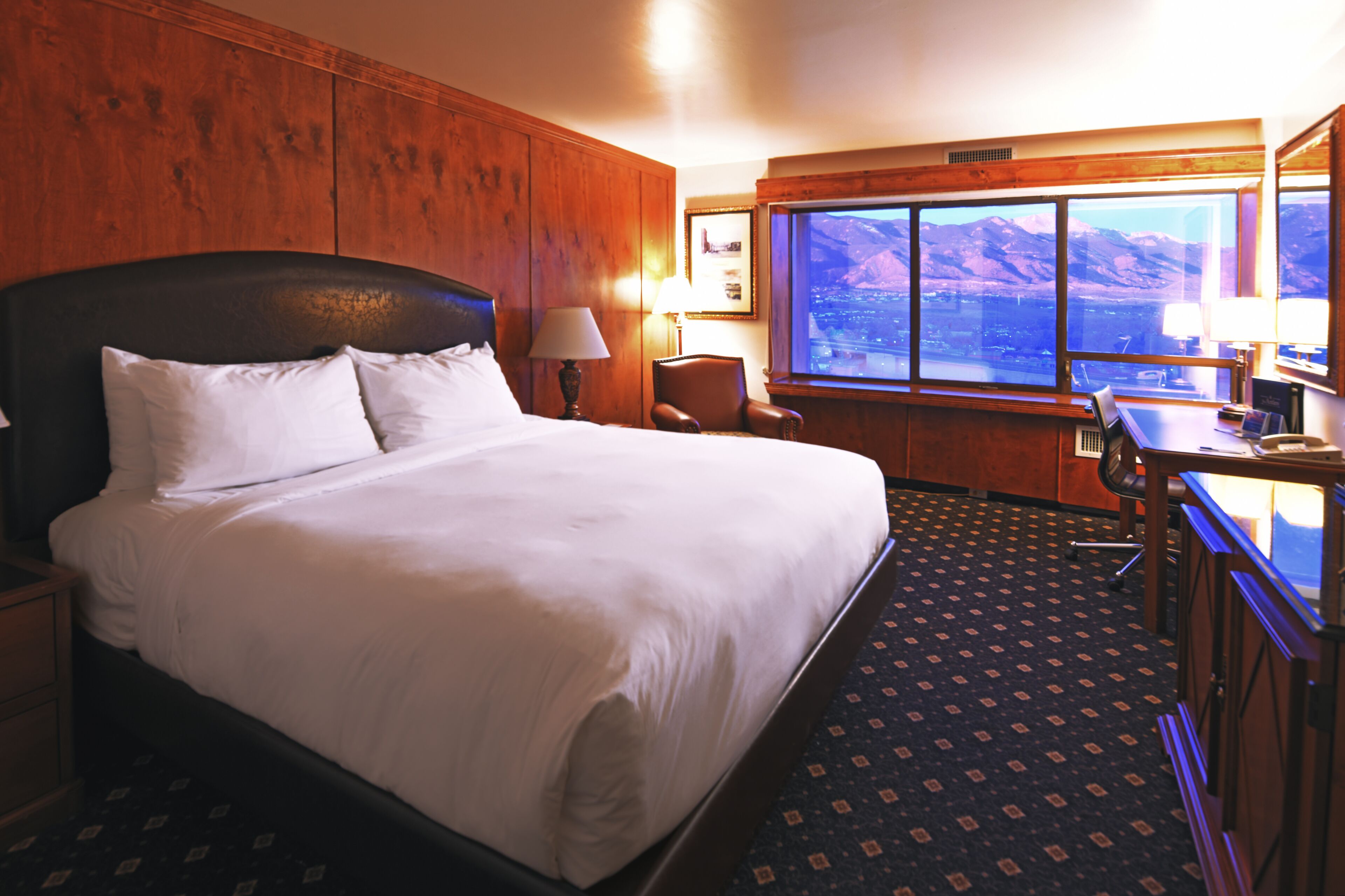A hotel room with a large bed, a desk, a chair, and a window with a mountain view.