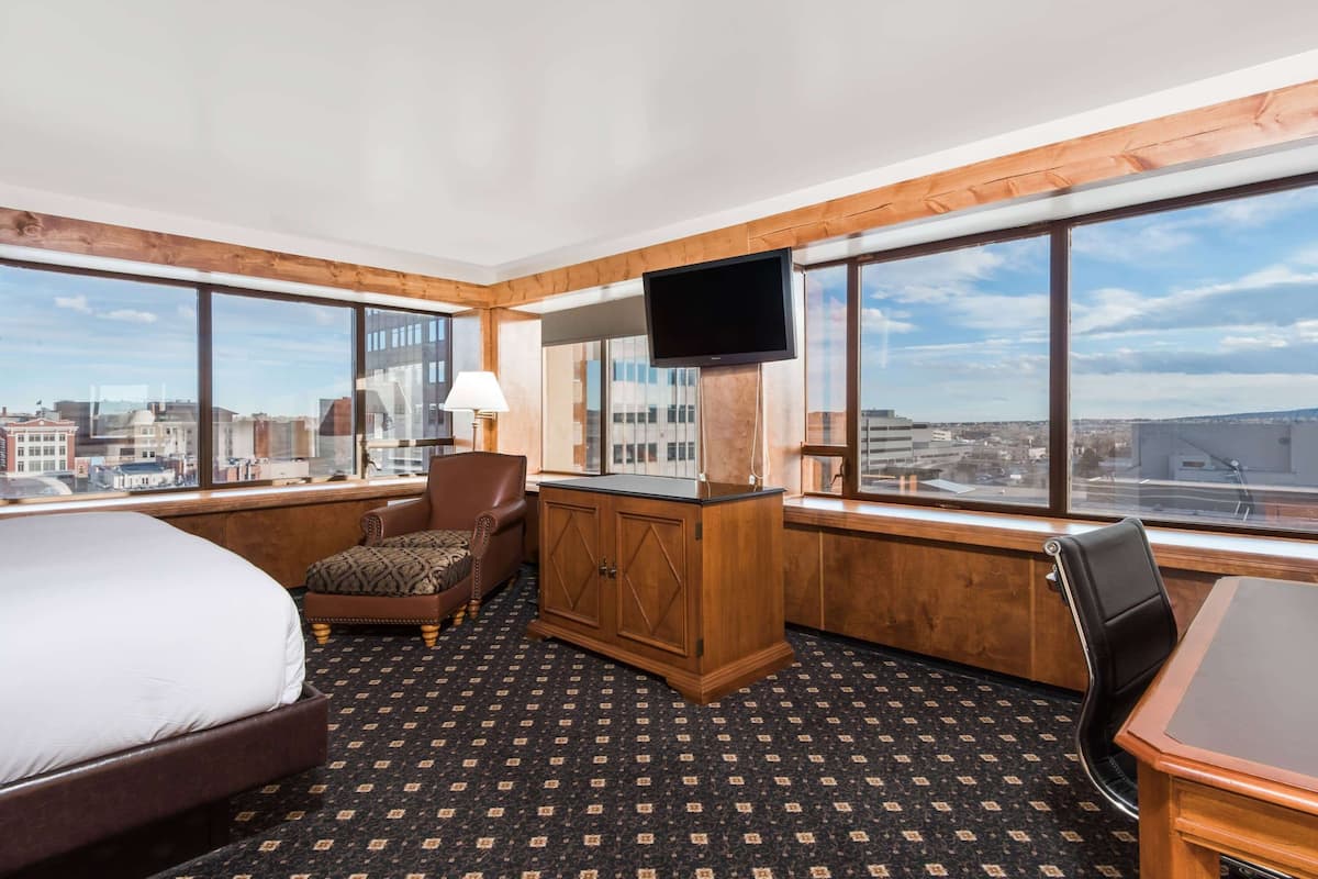 A hotel room with a large bed, a desk with a chair, a TV mounted on the wall, and large windows offering a city view.
