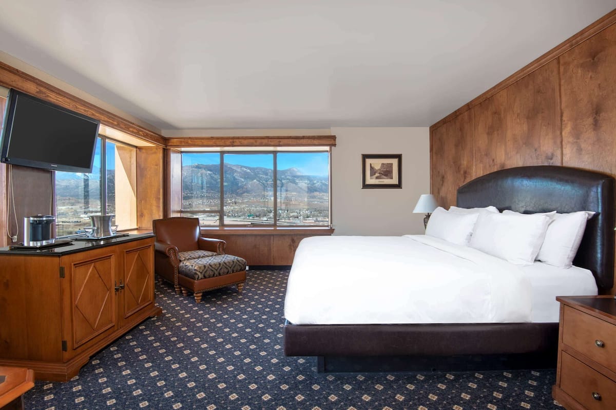 A hotel room with a large bed, a TV mounted on the wall, a wooden cabinet, a chair, and a window with a mountain view.