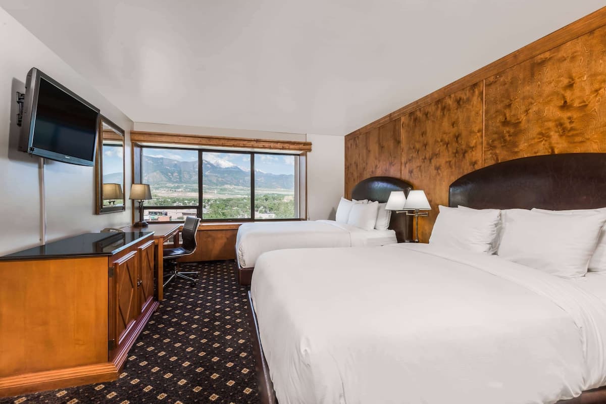 A hotel room with two beds, a desk, a TV, and a large window with a mountain view.