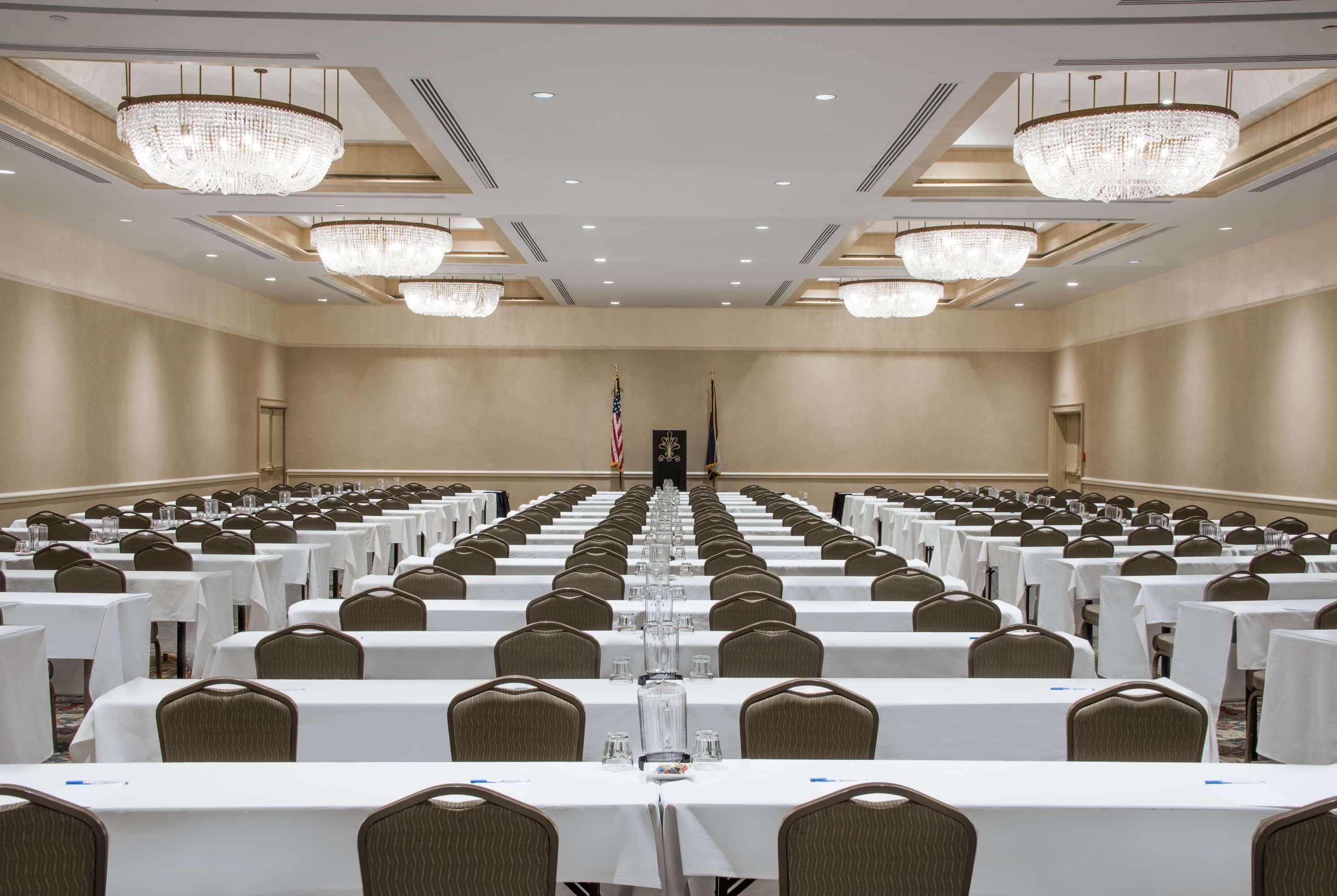 A conference room with rows of tables and chairs, chandeliers, and flags.