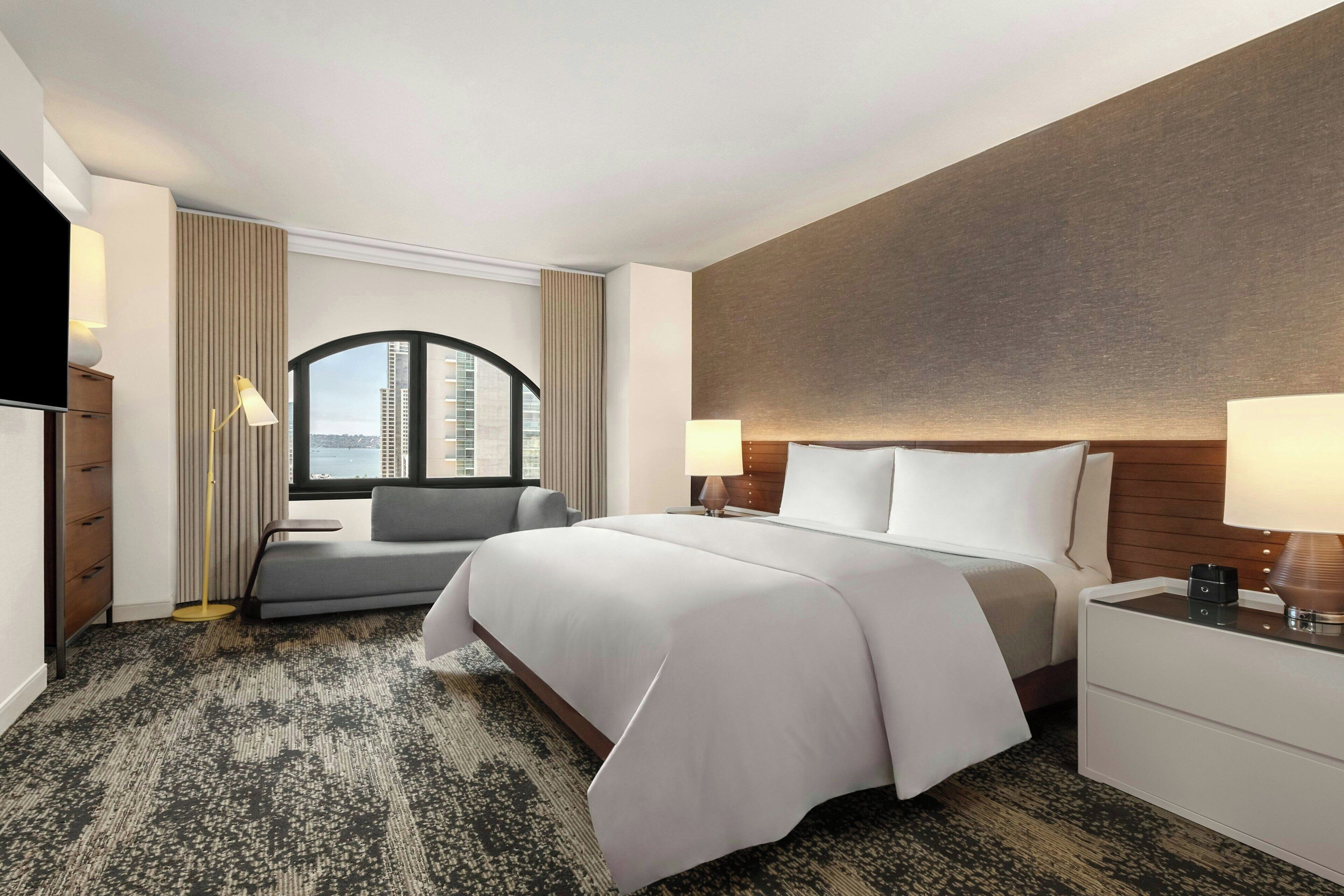 A hotel room with a large bed, a sofa, a desk, and a view of the city through a large window.