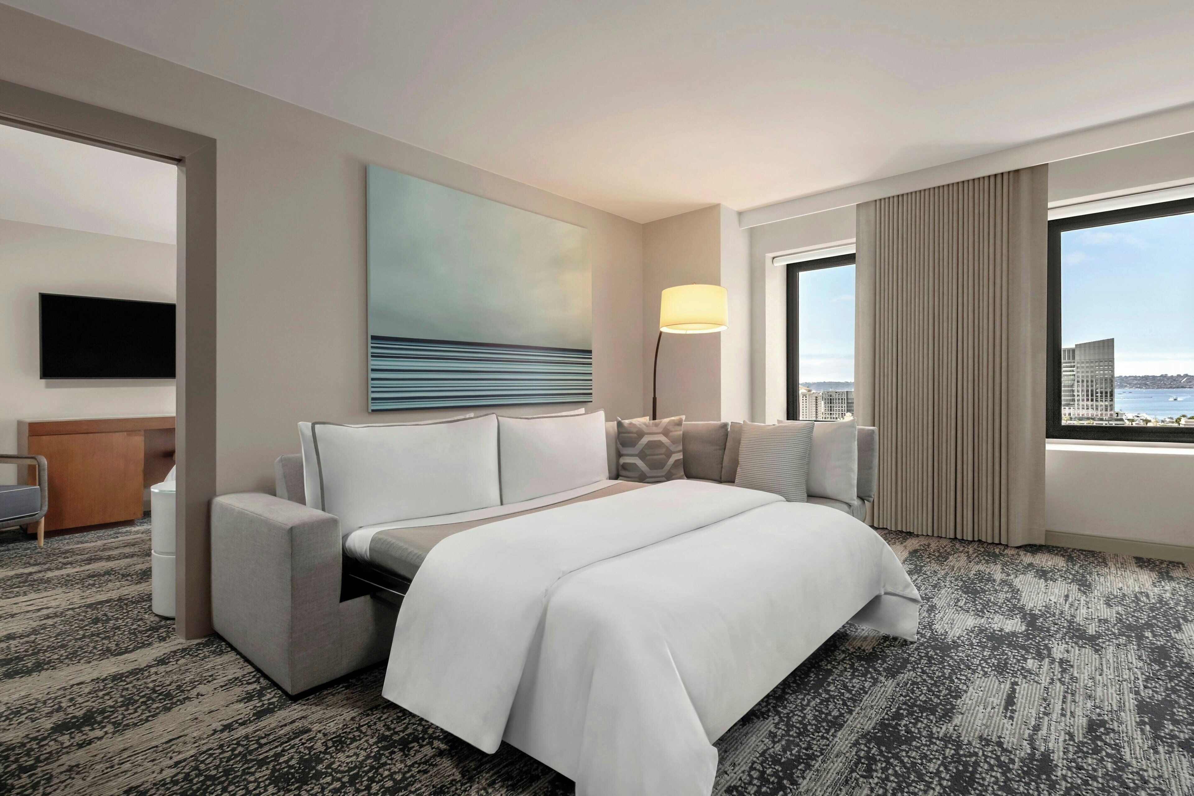 Ein modernes Hotelzimmer mit einem großen Bett, einem Flachbildfernseher, Blick auf die Stadt und einem großen Fenster mit Vorhängen.