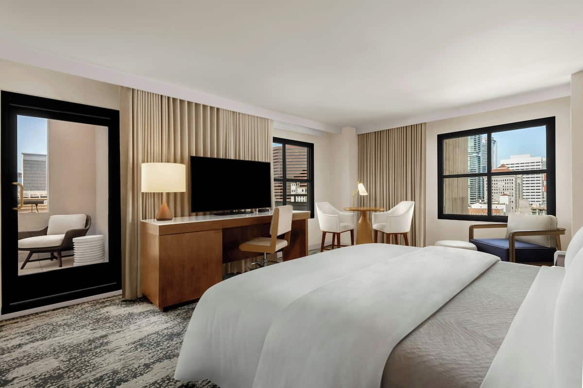 Habitación de hotel con una cama grande, un escritorio con televisor de pantalla plana, dos sillones, un sofá y vistas a la ciudad a través de amplias ventanas.