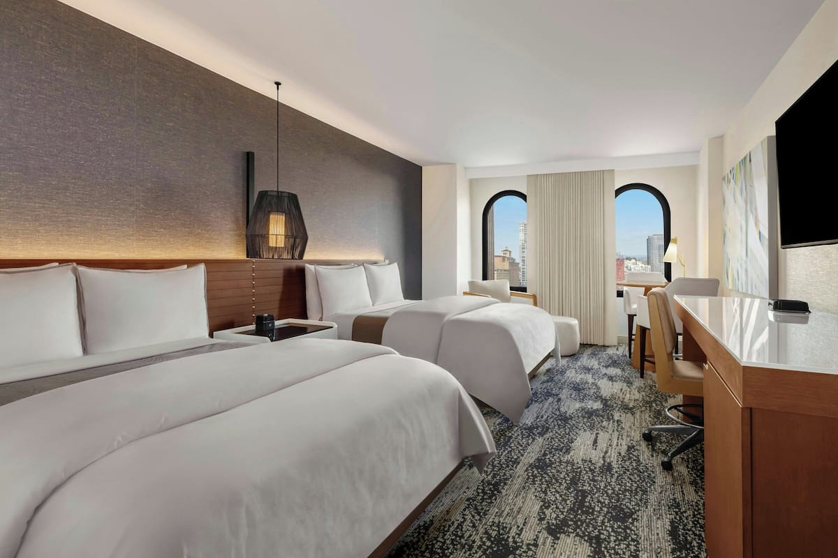 Habitación de hotel con dos camas, un escritorio y un ventanal con vistas a la ciudad.