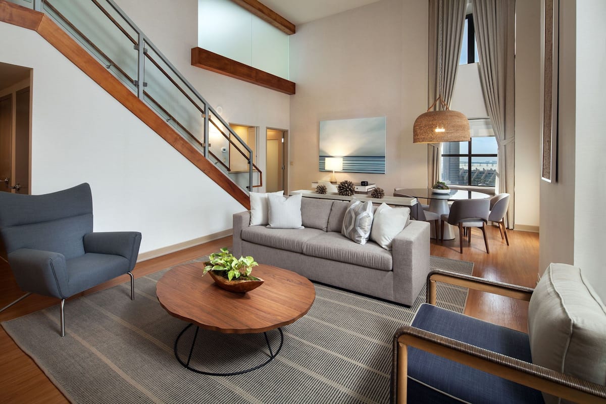 Una sala de estar moderna con un sofá gris, un sillón azul, una mesa de centro de madera y una escalera con barandillas de vidrio.