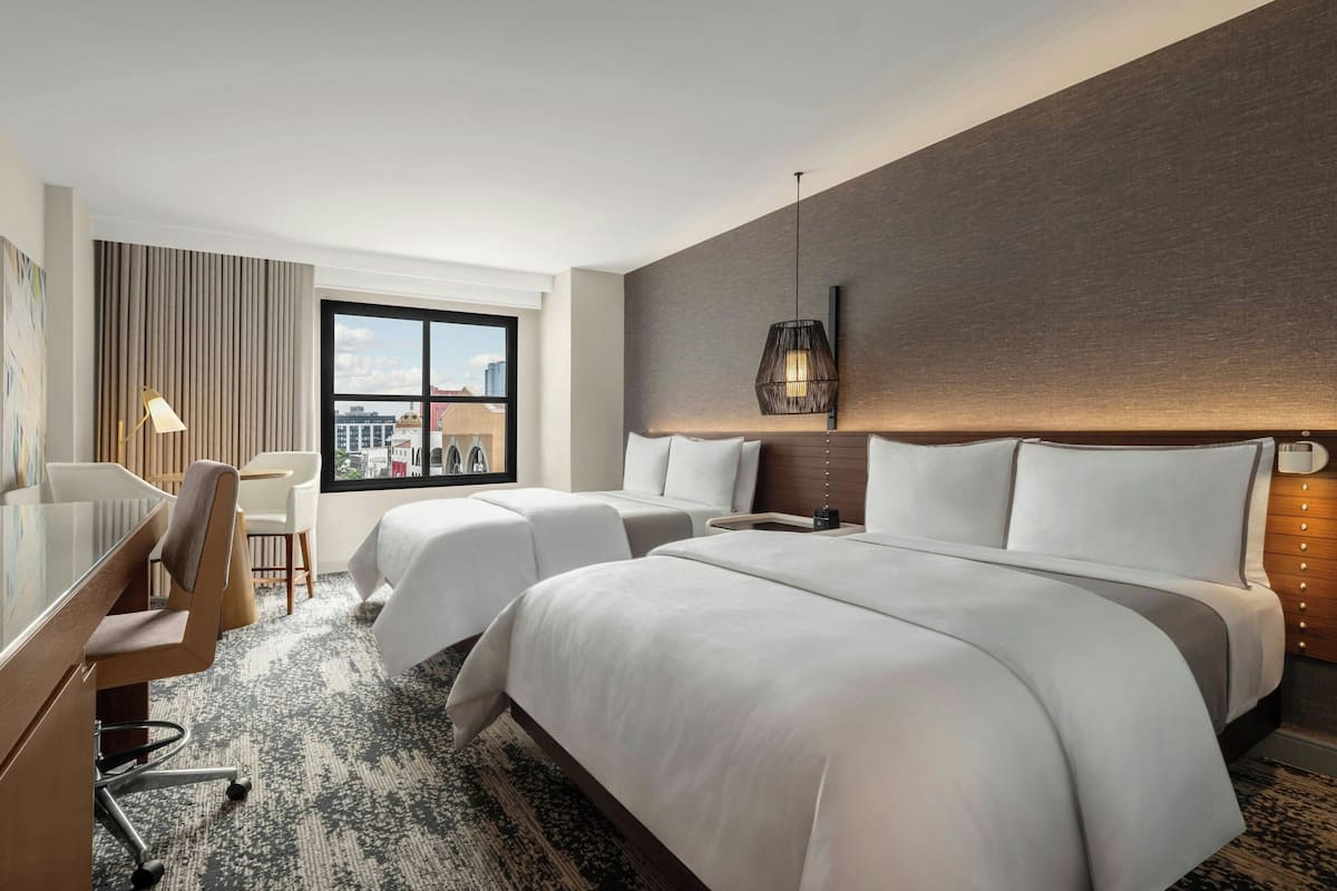 Habitación de hotel con dos camas, un escritorio, una silla, un ventanal grande con vistas a la ciudad y una lámpara colgante moderna.