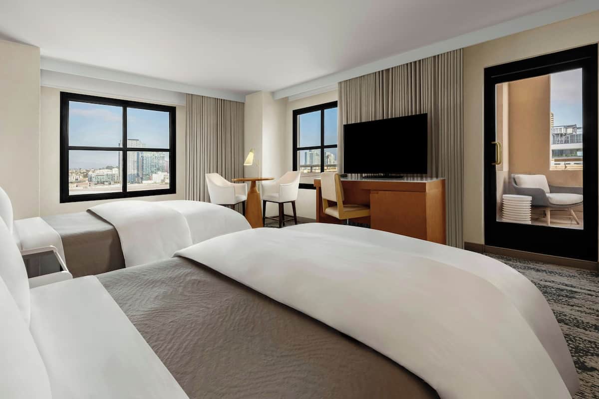 Habitación de hotel con dos camas, un escritorio con silla, televisión y amplias ventanas con vistas a la ciudad.