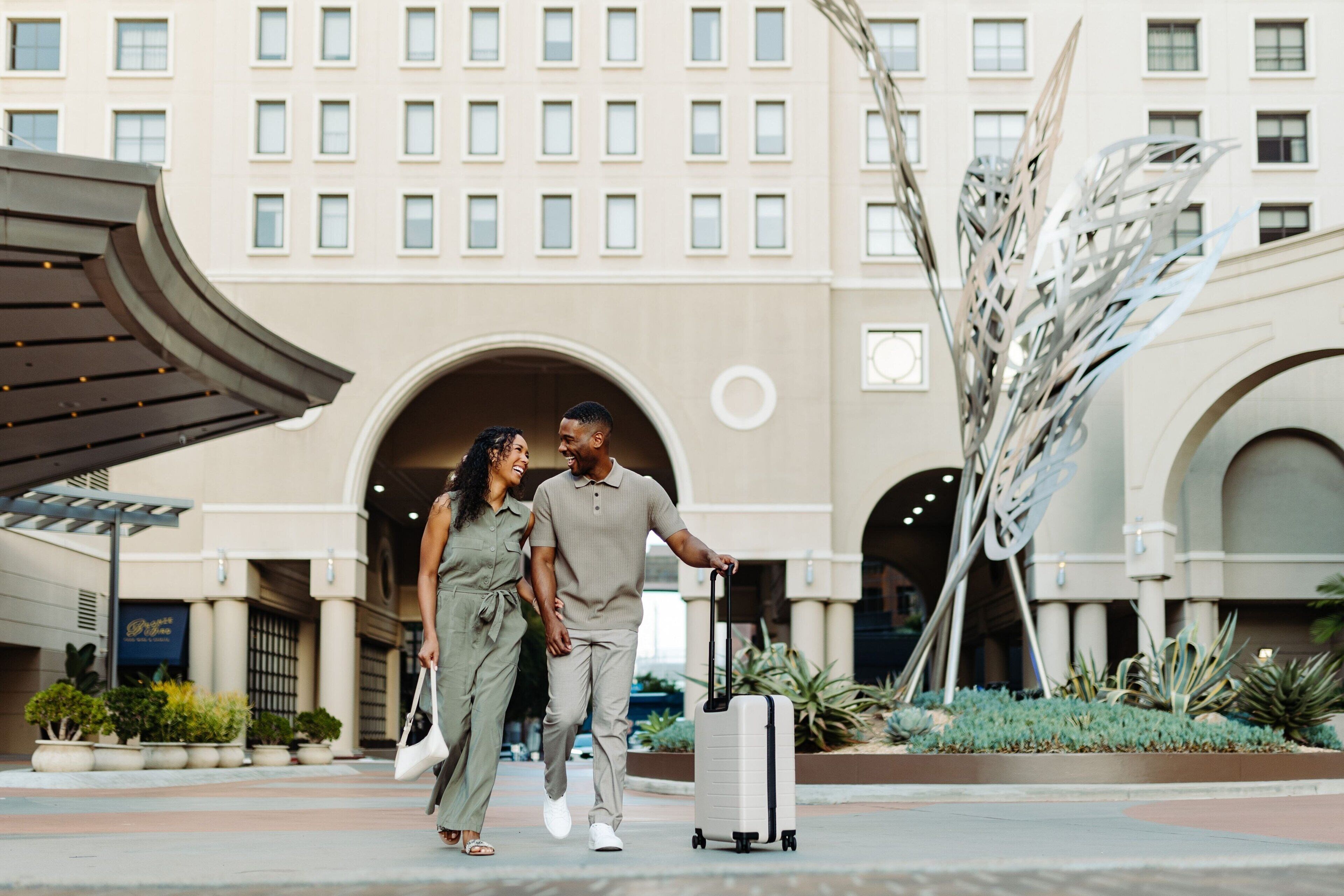 Una pareja caminando hacia la entrada de un hotel de diseño arquitectónico moderno, con arcos y una escultura decorativa de metal.